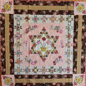 May include: A handmade quilt with a central star design featuring a floral arrangement in a yellow pot. The quilt has a pink background with floral and geometric patterns in brown, beige, and other colors. The quilt measures approximately 70 inches by 70 inches.