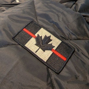 Può includere: Una toppa rettangolare con un disegno a foglia d'acero canadese. La toppa ha un bordo nero, uno sfondo grigio e bianco e una striscia rossa. La foglia d'acero è nera. La toppa è su un tessuto trapuntato nero.