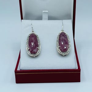 Puede incluir: Un par de pendientes de plata con grandes piedras preciosas rosas ovaladas y facetadas, rodeadas por un halo de pequeñas piedras transparentes. Los pendientes se exhiben en una caja de joyería roja con interior blanco.