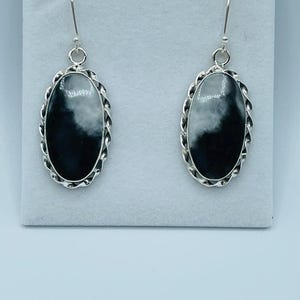 May include: A pair of oval earrings featuring a black and white marbled pattern. The earrings have a silver-coloured, twisted rope-style border and a simple hook fastening. They are presented against a white backdrop, showcasing their design.