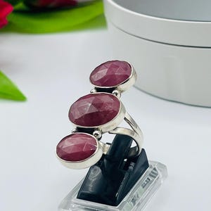 Könnte beinhalten: Ein silberner Ring mit drei ovalen rosafarbenen Edelsteinen. Der Ring befindet sich auf einem schwarzen Präsentationsständer.