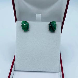 以下が含まれることがあります： 赤いジュエリーボックスに入ったエメラルドスタッドピアス。ピアスは、シルバーのセッティングが施された鮮やかなグリーンの宝石が特徴です。ボックスは開いており、柔らかい白い背景にピアスが展示されています。