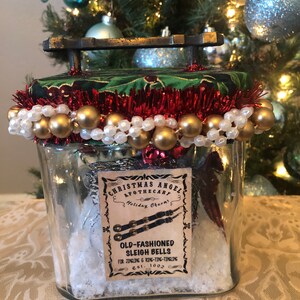Könnte beinhalten: Ein Glasgefäß mit einem roten und grünen Stoffdeckel, verziert mit goldenen Perlen und weißen Perlen. Das Gefäß enthält weißen Schnee und ein Etikett mit der Aufschrift "Christmas Angel Apothecary Holiday Charms Old-Fashioned Sleigh Bells for Jingling & Ring-Ting-Tingling Est. 1892".