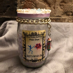 Könnte beinhalten: Ein Glasgefäß mit einem lila Deckel und einer goldenen Kette um den oberen Rand. Das Gefäß ist mit lila Glitzer gefüllt und trägt ein Etikett mit der Aufschrift "Bird Witch Apothecary Pixie Dust". Das Etikett zeigt auch eine Fee und den Text "Wesentlicher Bestandteil für Liebestränke und Anziehungssprüche. Est. 1692". Ein kleiner, bunter Kristall hängt an der Kette.