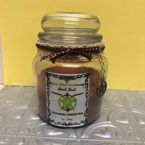 May include: A glass jar with a brown lid and a brown and white braided ribbon tied around the top. The jar is filled with a red and brown substance and has a label that reads "Burnt Witch Apothecary Devil Dust For summoning a crossroads demon Est. 1692".