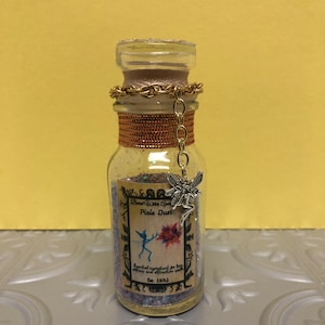 Op de afbeelding: Een klein glazen flesje met een kurkstop en een zilveren fee-bedel. Het flesje is gevuld met iriserende glitter en heeft een etiket met de tekst "Blessed With Pixie Dust. Essentieel ingrediënt voor liefdesdrankjes en aantrekkingsformules. Est. 1693."