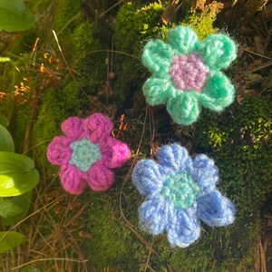 May include: Three crocheted flowers in different colors: pink, blue, and green. The flowers are all the same size and shape, with five petals and a small center. The flowers are arranged on a mossy surface.