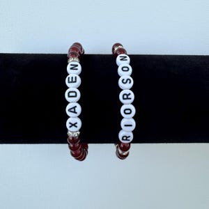 May include: Two stretch bracelets featuring deep red beads and white circular letter beads. One bracelet spells XADEN and the other spells RIORSON in black font. Both bracelets include small silver-toned accent spacers between the beads.