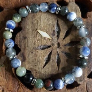 May include: A bracelet made of various blue, green, and grey stones with silver accents. The bracelet is laid out on a wooden surface.