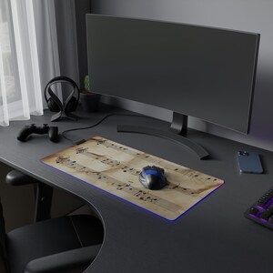 Peut inclure: Tapis de souris rectangulaire avec un motif de partitions musicales vintage et un bord lumineux bleu. Une souris d'ordinateur noire et bleue est posée sur le tapis. Le tapis est sur un bureau noir avec un écran d'ordinateur, un casque et une manette de jeu.