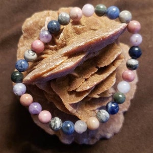 May include: A multi-coloured beaded bracelet with pink, blue and green stones. The bracelet is made of natural stones and is displayed on a brown rock.