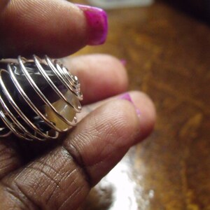 May include: A silver-colored wire cage pendant holding a combination of purple and yellow gemstones. The cage is spiraled around the stones, creating a unique design. The pendant is held in a hand with purple nail polish.
