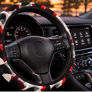 May include: A steering wheel cover featuring a repeating circular pattern in shades of red, black, white, and gray. The cover is installed on a black steering wheel inside a vehicle cabin, surrounding the central hub and spokes of the wheel.