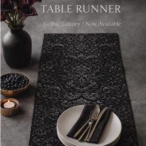 May include: A rectangular black table runner featuring a dark grey damask pattern. The text STYGIAN TABLE RUNNER and Gothic Luxury | Now Available is printed above the runner. A stack of white plates, a black napkin, and silver cutlery are placed on top.