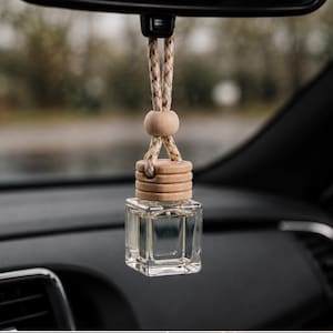 May include: A clear, square glass bottle hanging from a braided white and gold cord. The bottle features a light-colored wooden cap with horizontal grooves and a small, round wooden bead above it, designed for suspension from a vehicle mirror.