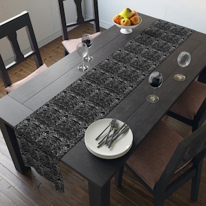 May include: A black table runner featuring an intricate, repeating heart-shaped filigree pattern with silver-toned highlights. The runner is laid across a dark wooden dining table set with white plates, silver forks, and clear glass wine goblets.