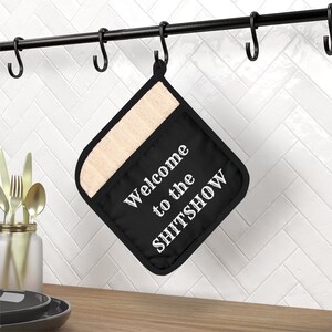 Puede incluir: Manopla de cocina negra con el texto blanco "Welcome to the SHITSHOW". Tiene un forro beige resistente al calor y cuelga de una barra de metal negro con ganchos. Utensilios de cocina.