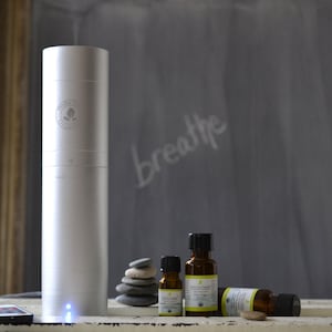 May include: A silver essential oil diffuser with a blue light, next to a stack of gray stones and three small amber glass bottles of essential oils. The bottles are labeled "100% Pure Organic" and "Breathe Easy Organic Citrus Sugar". The diffuser has a logo that says "Herbs & Earth Organics".