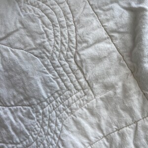 Peut inclure: Un tissu matelass&eacute; blanc pr&eacute;sentant une s&eacute;rie de lignes cousues parall&egrave;les et incurv&eacute;es qui cr&eacute;ent un motif textur&eacute; en forme de vagues sur toute la surface. Le mat&eacute;riau semble doux et mat, avec des d&eacute;tails de couture visibles partout.