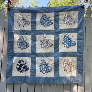 Peut inclure: Une courtepointe carr&eacute;e compos&eacute;e d'une grille de neuf blocs en tissu blanc cass&eacute;, chacun pr&eacute;sentant un papillon en tissu en relief avec divers motifs floraux ou &agrave; pois bleus, blancs et jaunes, bord&eacute;e d'un tissu bleu et blanc &agrave; micro-points.