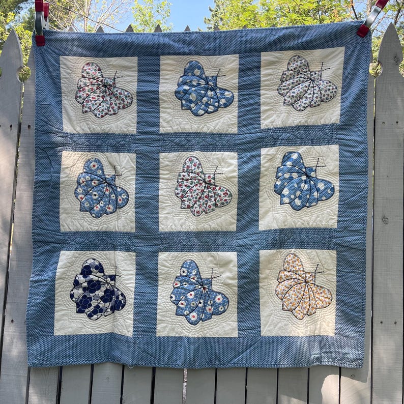 Peut inclure: Une courtepointe carr&eacute;e compos&eacute;e d'une grille de neuf blocs en tissu blanc cass&eacute;, chacun pr&eacute;sentant un papillon en tissu en relief avec divers motifs floraux ou &agrave; pois bleus, blancs et jaunes, bord&eacute;e d'un tissu bleu et blanc &agrave; micro-points.