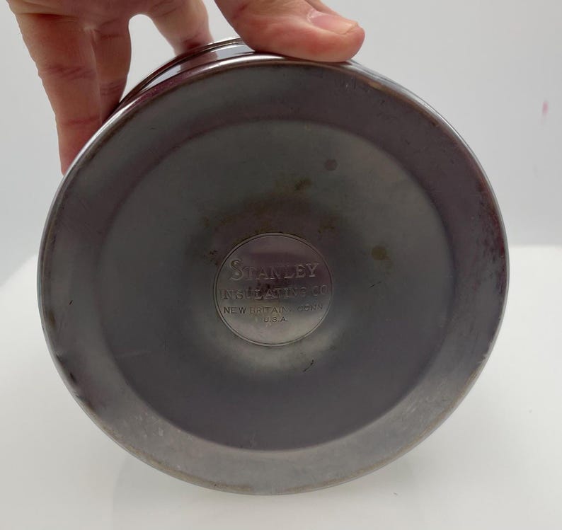 May include: A close-up of the base of a stainless steel Stanley vacuum flask. The base is a dark grey colour with a circular indentation. The Stanley logo is visible in the centre, with text that reads "Stanley Insulating Co. New Britain, Conn. U.S.A."