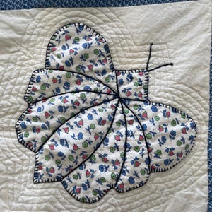Peut inclure: Un carr&eacute; de tissu blanc avec une application de papillon matelass&eacute;. Le papillon est en tissu blanc &agrave; petits motifs floraux bleus, roses et verts, soulign&eacute; par une &eacute;paisse couture noire. Deux fines antennes en fil noir d&eacute;passent du corps.