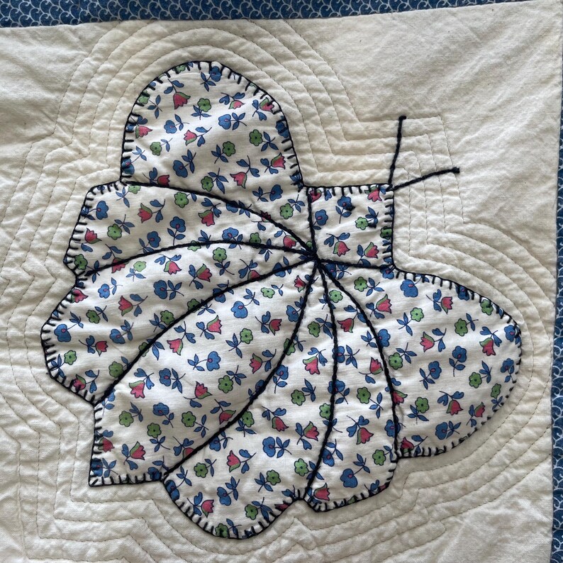 Peut inclure: Un carr&eacute; de tissu blanc avec une application de papillon matelass&eacute;. Le papillon est en tissu blanc &agrave; petits motifs floraux bleus, roses et verts, soulign&eacute; par une &eacute;paisse couture noire. Deux fines antennes en fil noir d&eacute;passent du corps.