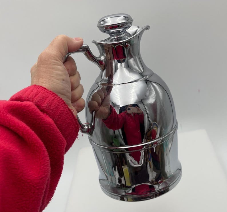 May include: A polished chrome jug with a handle and a lid. The jug has a rounded body and a slender neck. The surface reflects the surroundings. The jug is held by a person wearing a red sweater.