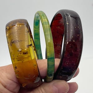 May include: Three resin bangles in shades of amber, green, and burgundy. The amber bangle has gold flecks, the green one has a marbled effect, and the burgundy one has gold glitter. The bangles are held in a hand.
