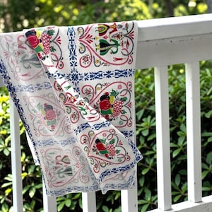 May include: A white tablecloth with a colorful floral and fruit pattern, including red grapes, green leaves, and blue accents. The tablecloth is draped over a white railing, showcasing its intricate design.