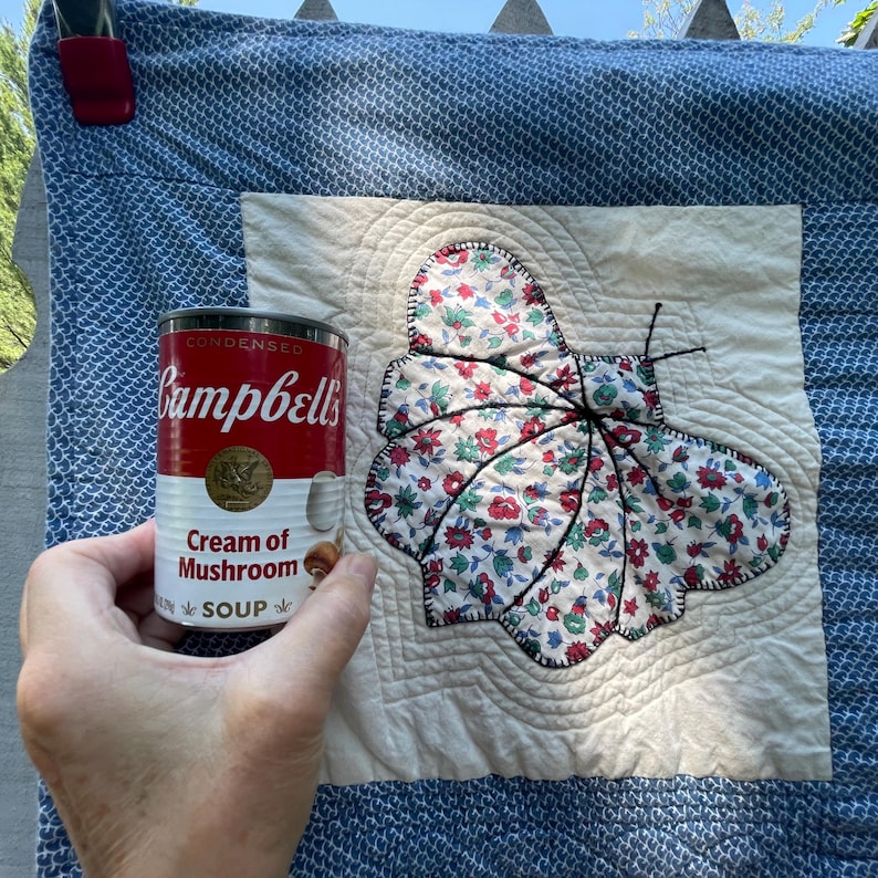 Peut inclure: Un carr&eacute; de courtepointe &agrave; motifs bleus et blancs avec un empi&egrave;cement central en tissu cr&egrave;me orn&eacute; d'un papillon floral en appliqu&eacute;. Une bo&icirc;te de soupe Campbell's Cream of Mushroom rouge et blanche est tenue devant pour l'&eacute;chelle.