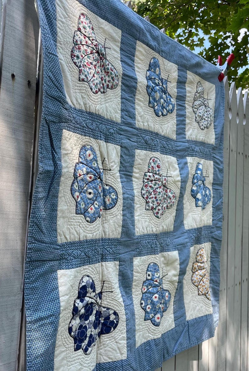 Peut inclure: Un quilt en patchwork compos&eacute; d'une grille de carr&eacute;s en tissu blanc cass&eacute;, chacun pr&eacute;sentant une applique de papillon unique faite de divers tissus &agrave; motifs bleus, blancs et floraux. Les carr&eacute;s sont s&eacute;par&eacute;s par des bordures en tissu textur&eacute;.