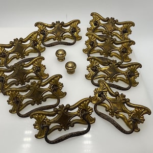 May include: A collection of antique brass drawer pulls and knobs. The drawer pulls have an ornate design with a central diamond shape and curved handles. The knobs are small and round. The hardware is a warm gold color.