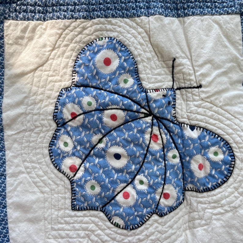 Peut inclure: Un bloc de courtepointe carr&eacute; pr&eacute;sentant une applique centrale en forme de papillon faite de tissu bleu &agrave; motif floral blanc avec de petits points rouges, verts et bleu fonc&eacute;. Le papillon est soulign&eacute; par des coutures noires sur un fond blanc.