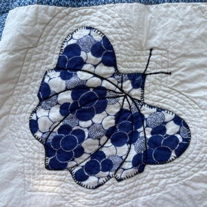 Peut inclure: Un carr&eacute; de courtepointe en tissu blanc avec une applique centrale en forme de papillon faite de tissu &agrave; motifs circulaires bleus et blancs. Le papillon est soulign&eacute; par des coutures noires et poss&egrave;de des antennes en fil noir sur un fond matelass&eacute;.