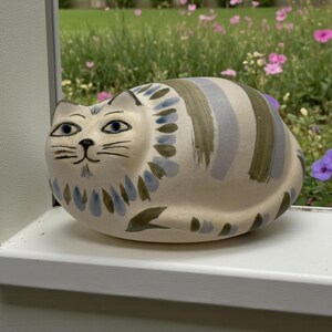 May include: A ceramic cat figurine with a cream-colored base and blue, green, and gray painted stripes. The cat has blue eyes and a detailed face. The figurine is sitting on a white windowsill, with a garden in the background.