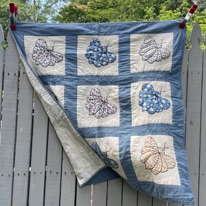 Peut inclure: Une courtepointe carr&eacute;e compos&eacute;e d'une grille de blocs en tissu blanc, chacun pr&eacute;sentant une applique de papillon unique dans divers motifs bleus, floraux et jaunes. La courtepointe est bord&eacute;e d'un tissu &agrave; micro-carreaux bleu et blanc.
