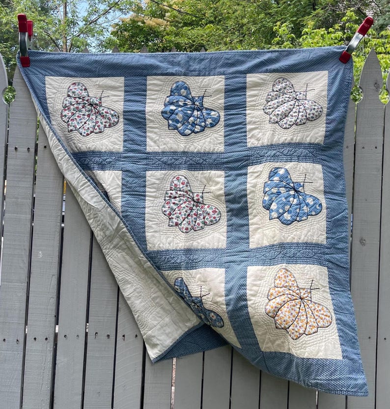 Peut inclure: Une courtepointe carr&eacute;e compos&eacute;e d'une grille de blocs en tissu blanc, chacun pr&eacute;sentant une applique de papillon unique dans divers motifs bleus, floraux et jaunes. La courtepointe est bord&eacute;e d'un tissu &agrave; micro-carreaux bleu et blanc.