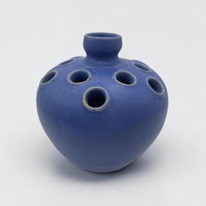 May include: A matte blue ceramic flower frog vase with a rounded, bulbous body and a central neck. The top surface features multiple circular openings arranged around the center, each with a light-colored rim, designed for holding individual stems.