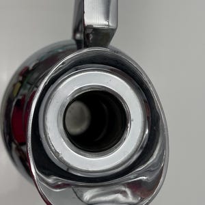 May include: Close-up of a shiny, chrome-plated flask with a handle. The flask has a wide opening with a silver rim and a dark interior. The handle is rectangular and also chrome-plated. The overall design is sleek and modern.