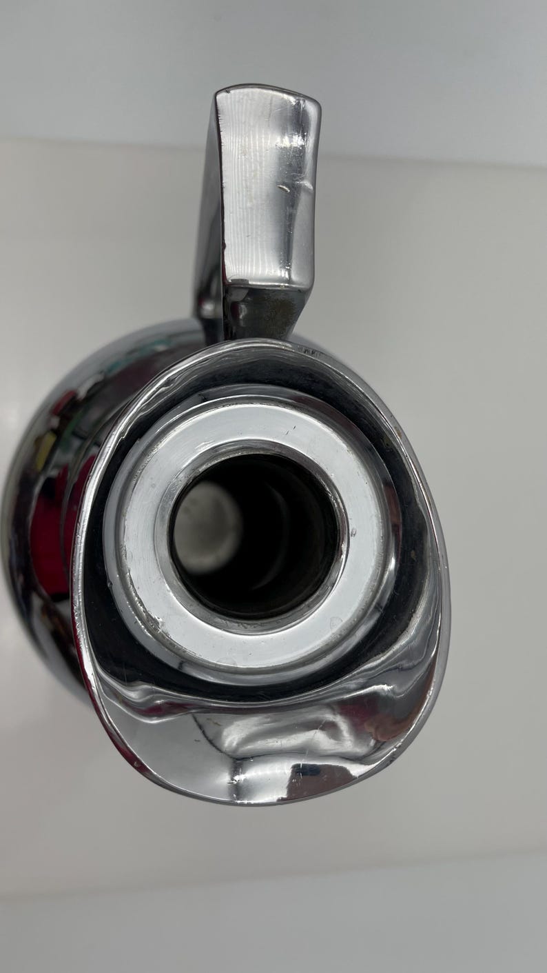 May include: Close-up of a shiny, chrome-plated flask with a handle. The flask has a wide opening with a silver rim and a dark interior. The handle is rectangular and also chrome-plated. The overall design is sleek and modern.