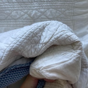 Peut inclure: Un textile matelass&eacute; blanc pr&eacute;sentant un motif de feuilles ou de p&eacute;tales cousu sur toute la surface. Le tissu a un aspect doux et textur&eacute; avec des coutures visibles. Une partie du bord r&eacute;v&egrave;le une bordure en tissu &agrave; motifs bleu et blanc.