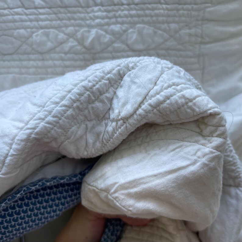 Peut inclure: Un textile matelass&eacute; blanc pr&eacute;sentant un motif de feuilles ou de p&eacute;tales cousu sur toute la surface. Le tissu a un aspect doux et textur&eacute; avec des coutures visibles. Une partie du bord r&eacute;v&egrave;le une bordure en tissu &agrave; motifs bleu et blanc.