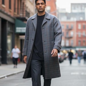 May include: A long, grey plaid wool-blend overcoat with a notched lapel, button-front closure, and cuffed sleeves, worn over a plain black t-shirt and dark grey trousers. The coat features a subtle brown and grey check pattern throughout the fabric.
