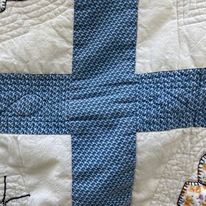 Peut inclure: Un textile matelass&eacute; pr&eacute;sentant un motif en forme de croix en tissu bleu avec un motif de vagues r&eacute;p&eacute;titif blanc. Les sections environnantes sont en tissu blanc cass&eacute; uni avec des lignes de matelassage courbes et des accents floraux.