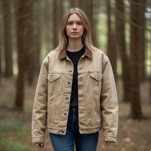 May include: A tan work jacket with a brown corduroy collar, worn over a black t-shirt and blue jeans. The jacket has button closures, two chest pockets, and a small red logo on the chest. The background is a blurred forest scene.