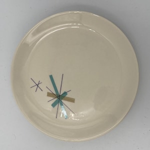 May include: Cream-colored ceramic plate with a mid-century modern design. The plate features a cluster of geometric shapes in teal, tan, and silver, with black lines radiating outward. A simple, star-like design is also present.