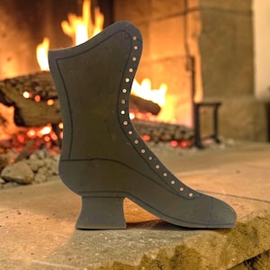 May include: A black decorative boot silhouette with copper-colored rivet details. The boot is placed on a stone surface in front of a fireplace with a burning fire. The boot has a high heel and a pointed toe.