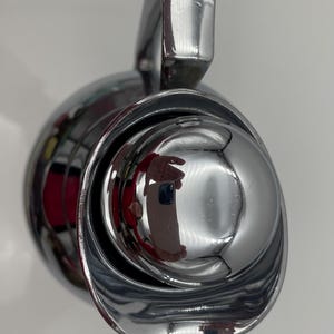 May include: A close-up of a shiny, chrome-plated object with a rounded, bowl-like shape and a curved handle. The surface reflects the surrounding environment, creating a distorted mirror effect. The object appears to be a faucet or similar fixture.